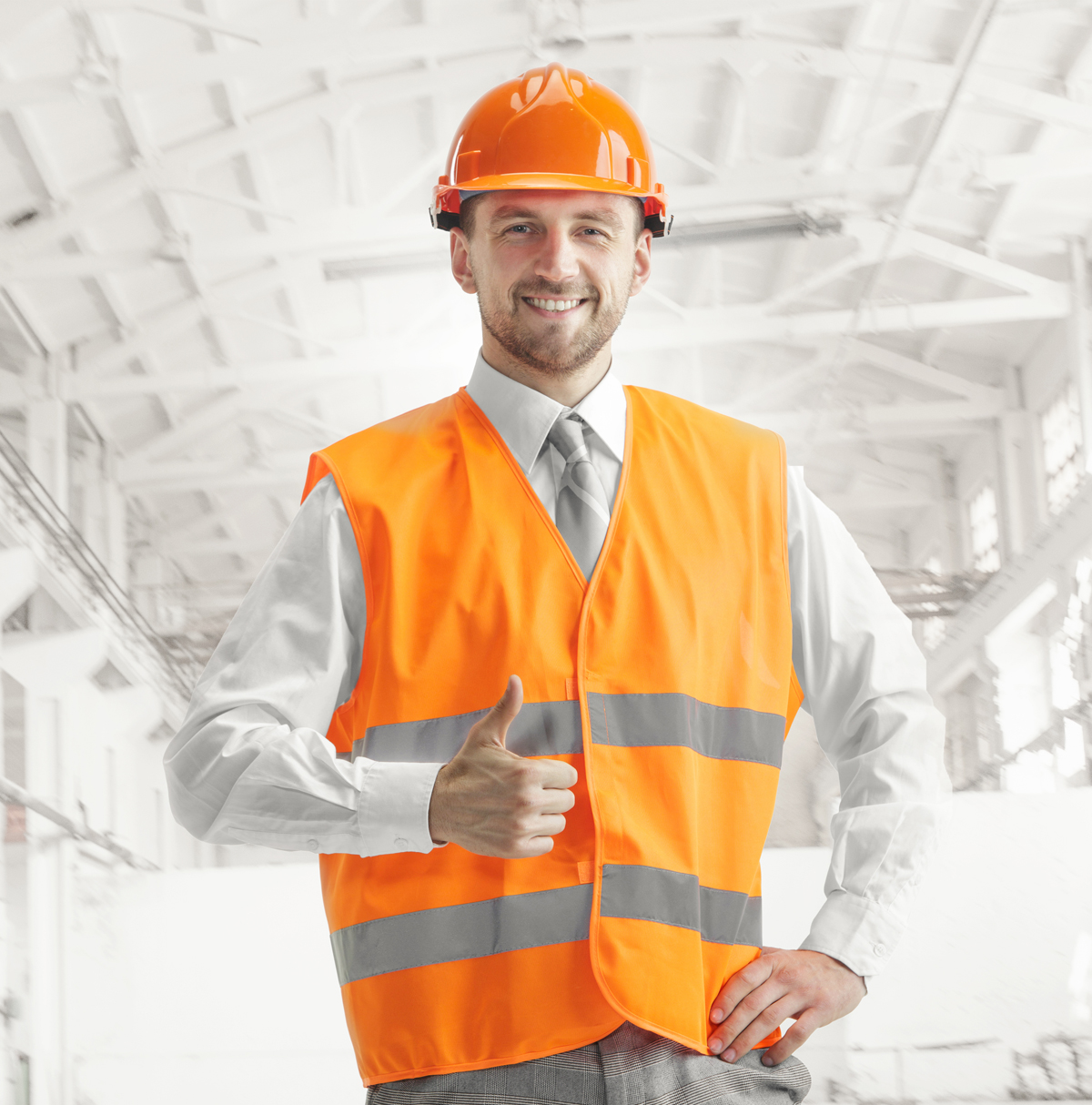 The builder in a construction vest and orange helmet smiling with sign ok against industrial background. Safety specialist, engineer, industry, architecture, manager, occupation, businessman, job concept
